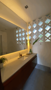 2024 Special Promo, A modern bathroom in the King Garden Room at Amila Dive Beach Resort features a long countertop and a rectangular sink. A backlit mirror hangs above, with a potted plant and toiletries on the counter. The wall boasts decorative geometric cutouts, allowing natural light to filter through.