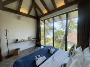 A bright King Birdview Room at Amila Dive Beach Resort features a large bed with white and navy blue bedding, facing a wall-sized window with tall curtains. Outside, palm trees and a glimpse of the ocean can be seen. The room includes a small desk, chair, air conditioning unit, coat rack, and various amenities.