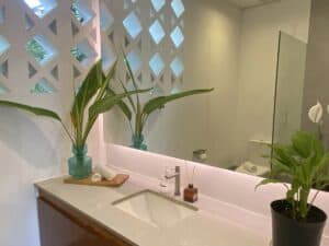 A modern bathroom in the King Garden Room at Amila Dive Beach Resort features a geometric lattice wall, a large mirror, and a white sink on a light countertop. The counter holds a teal vase with large leaves, a soap dispenser, and a small plant. A black potted plant is placed on the right side of the counter.