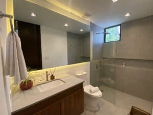 A modern bathroom in the Superior King Room at Amila Dive Beach Resort features a large mirror, under-cabinet lighting, and a sink on a wooden vanity. A towel hangs on a rod to the left, and a glass-enclosed shower with a small window is on the right. Decorative items and a plant enhance the vanity's elegance.