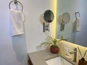 A Superior King Room at Amila Dive Beach Resort boasts a modern bathroom with a wall-mounted towel ring holding a white towel, an extendable shaving mirror, and a countertop featuring a sink, soap dispenser, potted plant in a red pot, and brown box. The walls and mirror are bathed in warm light.