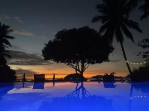 A serene tropical sunset with a glowing blue infinity pool in the foreground at Amila Dive Beach Resort. Silhouettes of palm trees and lounge chairs border the pool, while string lights add a cozy ambiance. The sky is painted in shades of orange and purple, reflecting on the water's surface.