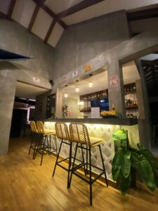 A cozy bar with high ceilings and warm lighting features a counter with four wicker barstools. The bar, part of the Restaurant Langga at Amila Dive Beach Resort, is illuminated from below, and bottles and decor line the shelves behind. Green plants add a touch of nature to the modern, concrete interior.