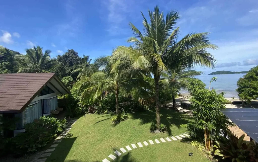 A tropical beachfront scene at Amila Dive Beach Resort features a small cottage on the left, surrounded by lush greenery and palm trees. A stone path winds through the neatly manicured lawn towards the ocean, which is visible in the background beneath a clear, blue sky.