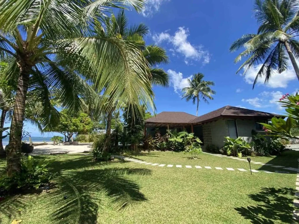 A tropical scene featuring a cozy Superior King Room bungalow surrounded by lush green lawns and various palm trees. The clear blue sky and ocean in the background enhance the serene atmosphere, while a stone pathway leads up to the inviting bungalow.