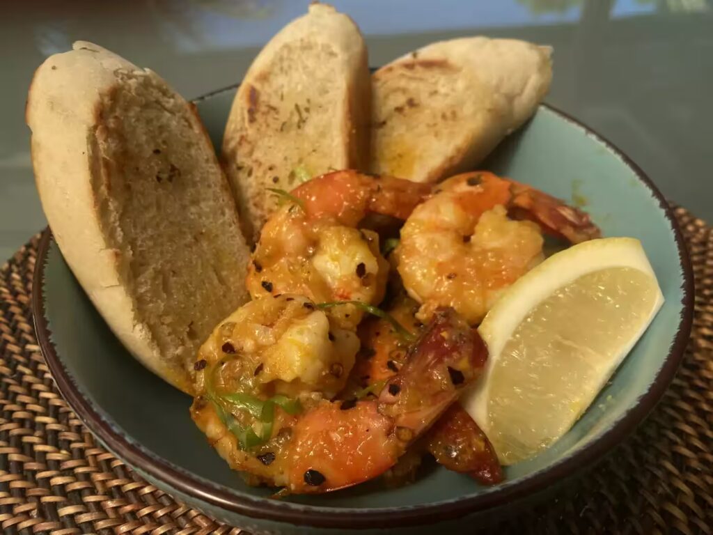 A Bowl Of Seasoned Shrimp Garnished With Herbs, Accompanied By Three Slices Of Toasted Bread And A Lemon Wedge, Placed On A Round Woven Mat.