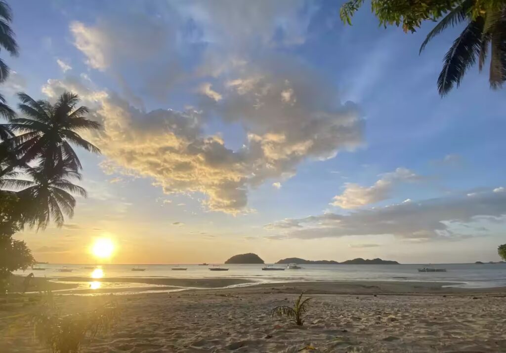 Sunset Over A Tranquil Beach With Golden Skies And Scattered Clouds. Silhouettes Of Palm Trees Frame The Image, And Small Boats Are Visible On The Horizon, Beneath A Vibrant Sky Reflecting On Calm Waters.