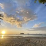 Sunset Over A Tranquil Beach With Golden Skies And Scattered Clouds. Silhouettes Of Palm Trees Frame The Image, And Small Boats Are Visible On The Horizon, Beneath A Vibrant Sky Reflecting On Calm Waters.